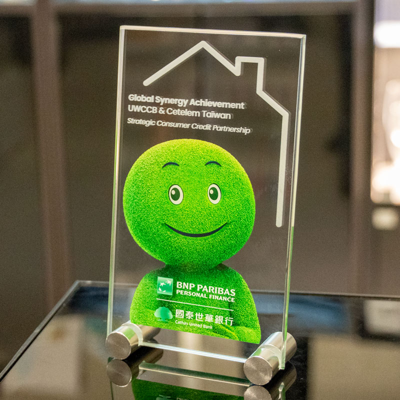 Plaque en verre verticale sur deux pieds métalliques, personnalisée avec la mascotte verte Cetelem et des logos en couleur.