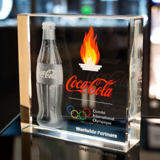 Trophée en verre avec bouteille Coca-Cola gravée en 3D, logo rouge, flamme et anneaux olympiques en couleur.
