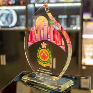 Trophée en verre en forme de flamme sur socle, avec une ville en couleur, l’emblème des pompiers de Hong Kong et une dédicace.