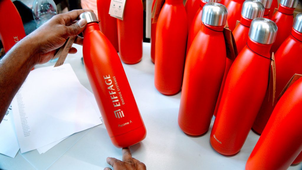 Gourde rouge tenue à la main, gravée du logo Eiffage Énergie Systèmes et d’un prénom, devant une série de bouteilles.v